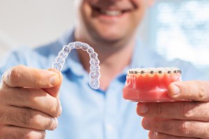 A smiling man holds a model of human teeth with braces and an Invisalign tray